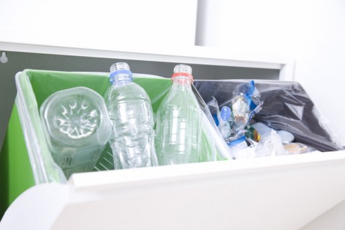 A close-up view of a recycling bin with a bright green interior, positioned within a clean, white environment, likely on a countertop or inside a waste collection area. Inside the bin are several clear plastic bottles, some with coloured caps—one blue and another red—showing various sizes and shapes of recyclable containers. To the right of the green bin is a black plastic bag filled with miscellaneous waste, including visible plastic items and packaging materials. The scene is well-lit, emphasizing the transparency of the plastic bottles and the contrasting colours of the caps and waste materials. The setting appears tidy and organized, representative of waste collection or rubbish removal services in Bounds Green, suitable for a professional rubbish removal company like Commercial Waste Removal Bounds Green, servicing local commercial and residential clients with environmentally conscious waste collection solutions.
