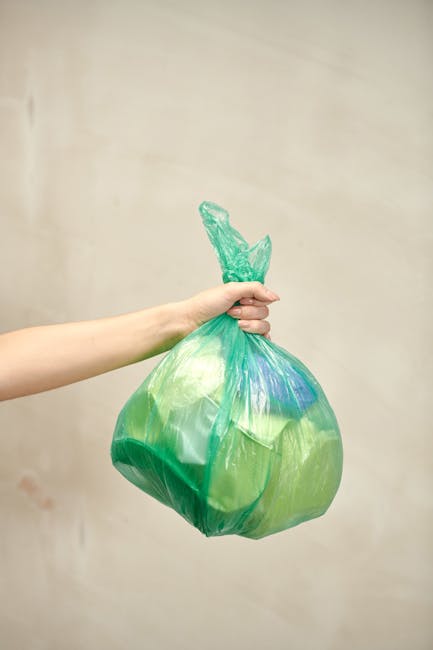 A green industrial waste bin filled with several red and black bags of garden or building material, placed outdoors against a dark brick wall. The bags are labeled with white text, indicating they con