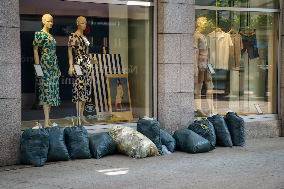 A person standing outdoors on a grassy area, wearing a plaid flannel shirt over a green top and grey trousers, holding an open black rubbish bag with green disposable gloves. The black plastic bag app